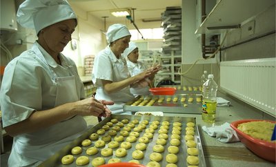 Two women wearing white uniforms, aprons, and chef's hats stand at a counter rolling several dozen cookies on trays